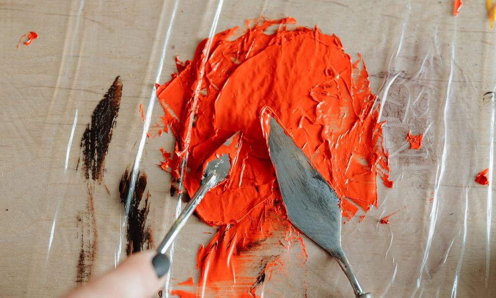 A person with dark nail polish mixes bright orange thick paint with a small palette knife. The paint is placed on transparent plastic sheet on a light colored wooden table.There is also a larger palette knife placed in the paint.
