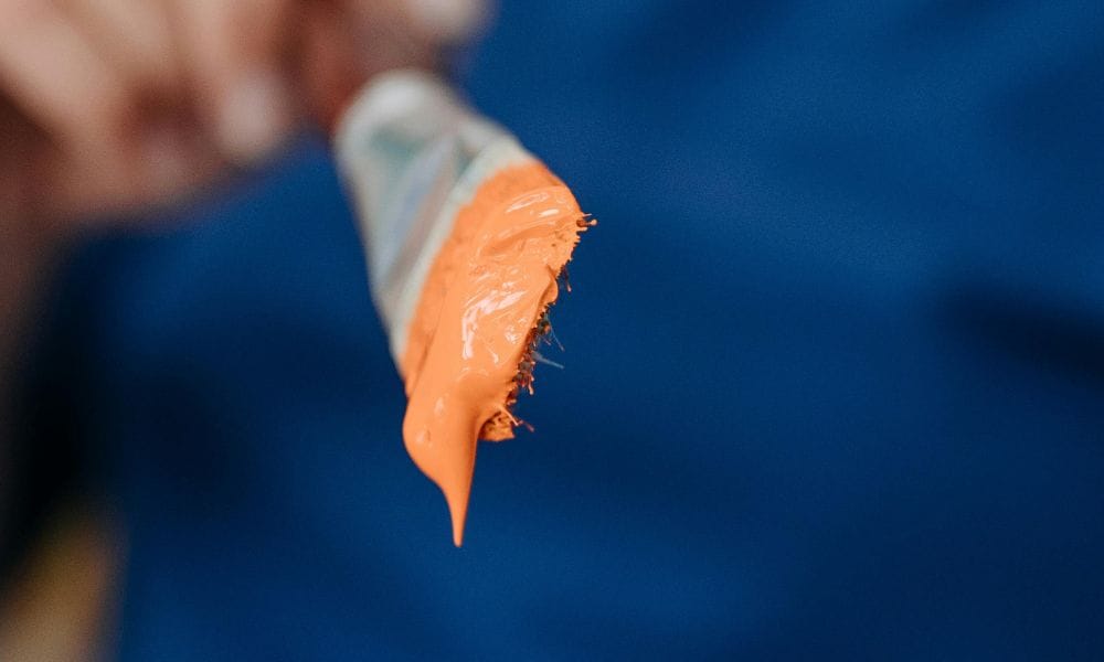a person holds up a flat paintbrush full of light orange paint towards the camera. the background is blurred. The person is wearing a blue shirt.